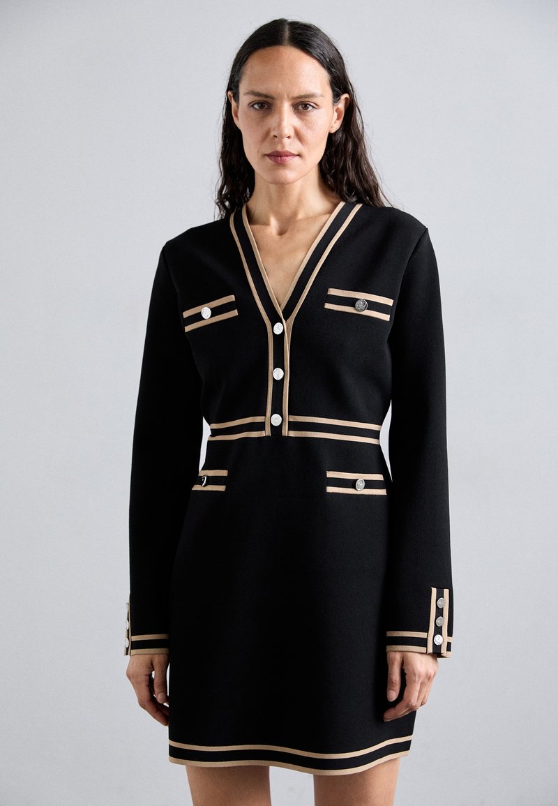 Robe noire à manches longues, décolleté en V, rayures crème, et boutons ronds en argent sur les poches et les poignets. Tissu lisse avec un design structuré.