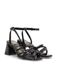 Zapatos de charol negro con tacón cuadrado, que cuentan con múltiples tiras entrelazadas y una correa en el tobillo. Plantilla texturizada con letras doradas.