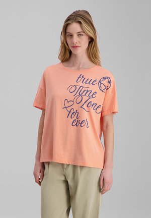 Junge Frau mit langen Haaren, die ein pfirsichfarbenes T-Shirt mit blauem Text „true Time Zone for ever“ und einer kleinen Globusgrafik trägt, kombiniert mit beigen Hosen.