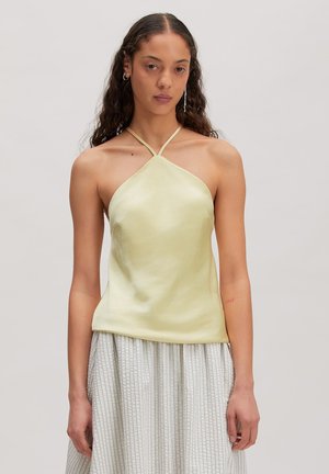Woman with curly hair wearing a pale yellow halter top and high-waisted, light-striped skirt standing against a plain background.