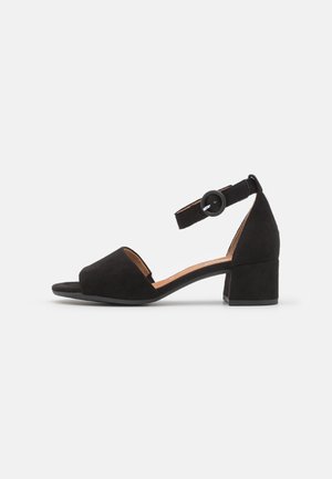 Sort ruskind åben-tå sandal med blokhæl og justerbar ankelrem med rund sort spænde, vist i sideprofil.