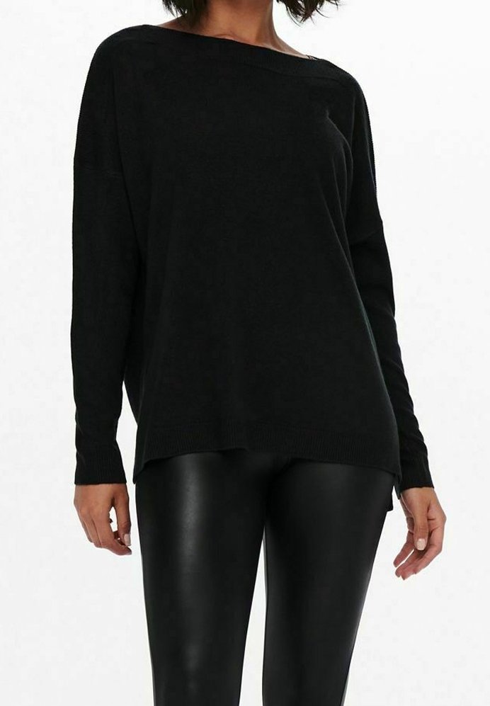 Femme portant un pull large noir à épaules dénudées et des leggings effet cuir noirs, debout devant un fond blanc uni.