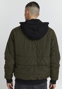 Giacca a vento verde oliva con cappuccio nero, texture a rombi, polsini a contrasto e vestibilità rilassata, vista da dietro.