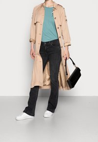 Beige trenčkot, tyrkysové tričko, černé džíny a bílé tenisky. Drží černou kabelku se zlatými detaily. Hladké materiály, ležérní střih.