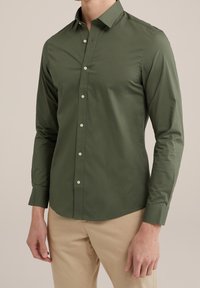 Mężczyzna w dopasowanej oliwkowej koszuli z długim rękawem z guziczkami oraz beżowych spodniach, stojący na tle jasnego, jednolitego tła.