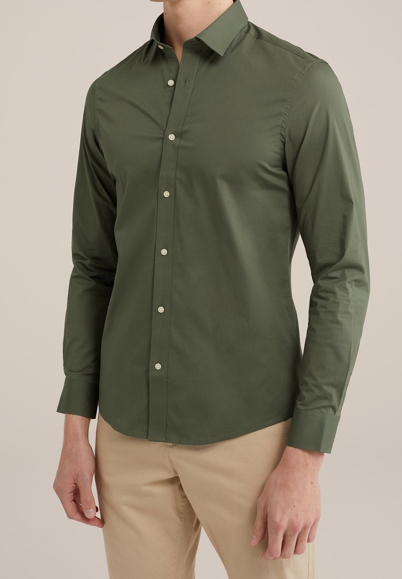 Mężczyzna w dopasowanej oliwkowej koszuli z długim rękawem z guziczkami oraz beżowych spodniach, stojący na tle jasnego, jednolitego tła.