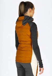 Gilet isolante arancione con cappuccio e accenti grigi, caratterizzato da una texture trapuntata e una vestibilità slim. Abbinato a un top scuro a maniche lunghe.