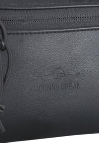 Schwarze strukturierte Tasche mit einem Reißverschluss. Mit geprägtem "JOHNNY URBAN"-Logo und einem dekorativen Kordeldetail an der Seite.