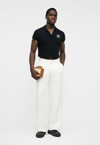 Homme portant un polo noir court, pantalon blanc à jambes larges, chaussures noires, tenant un petit sac à main marron, debout devant un fond clair uni.