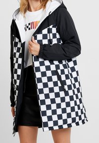 Kvinna som bär en svart-och-vit rutiga huoodjakke över en vit kortärmad grafisk t-shirt och en svart kjol.