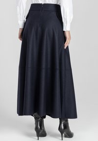 Kobieta w długiej, ciemnoniebieskiej spódnicy z wysokim stanem, połączonej z czarnymi botkami na obcasie i białą koszulą z długim rękawem.