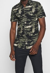 Kortärmelig overhemd met een zwart-groen camouflagepatroon. Gemaakt van lichtgewicht stof, met een knoopsluiting aan de voorkant en een kraag.