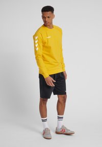 Geel sweatshirt met witte branding en chevronaccenten op de mouwen, gecombineerd met zwarte shorts en grijze sneakers met rode strepen.