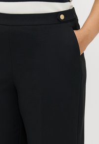 Mustat housut, joissa on sileä pinta, varustettuna kultaisella nappikoristeella vyötärön lähellä ja sivutaskuilla lantion alueella.