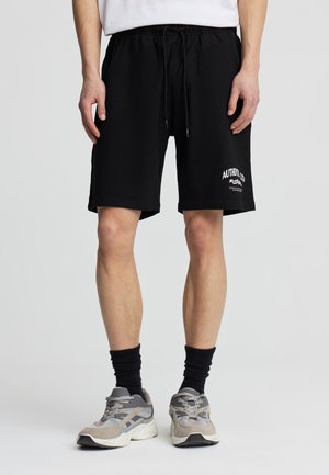 Gambe maschili che indossano pantaloncini neri con coulisse e logo bianco sulla gamba sinistra, calzini neri e scarpe da ginnastica grigie, in piedi su sfondo neutro.