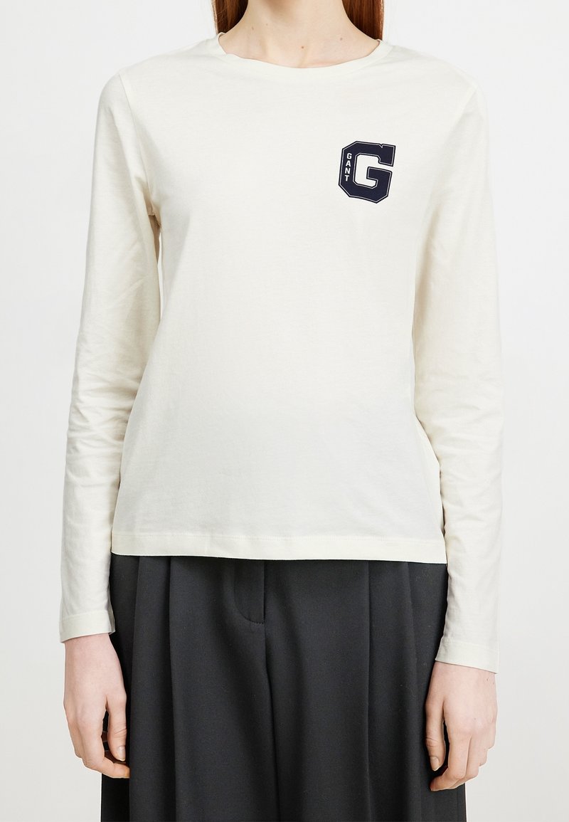 Langärmliges T-Shirt aus cremefarbener Baumwolle mit rundem Halsausschnitt, ausgestattet mit einem navyfarbenen GANT-Logo auf der Brust und einem lockeren Schnitt.
