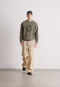 Olivovo zelená mikina s okrúhlym logom, spárovaná s béžovými nohavicami s širokými nohavicami a čiernymi topánkami. Model má na hlave čiernu čiapku s bielymi detailmi.