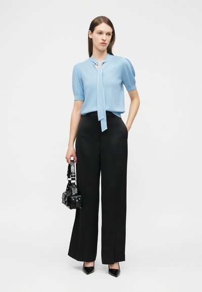 Femme portant un chemisier bleu clair à manches courtes et un pantalon noir à jambes larges, tenant un sac à main noir, se tenant devant un fond blanc.