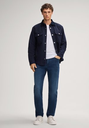 Marineblauw overhemdjacket met twee borstzakken en zilveren drukknoopsluitingen, gedragen over een wit T-shirt, gecombineerd met blauwe jeans en witte sneakers.