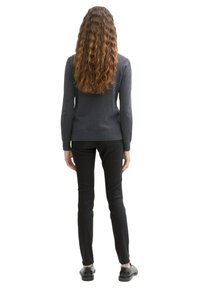 Grauer gestrickter Pullover mit langen Ärmeln, kombiniert mit schwarzen engen Hosen und schwarzen Schuhen, von hinten gesehen. Texturiertes Haar fällt unter die Schultern.