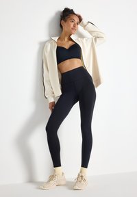 Musta urheiluliivi, kerma värinen vetoketjullinen takki, korkeavyötäröiset mustat leggingsit ja vaaleanbeige kengät, joissa on vaaleankeltaiset nilkkasukat. Sileä, tiukka kangas.