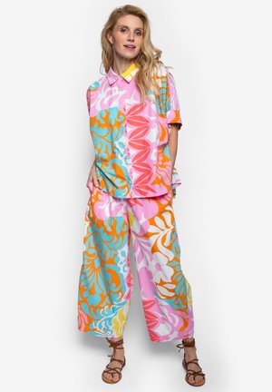 Kobieta ubraną w luźną, kolorową koszulę w kwiatowy wzór oraz szerokie spodnie w kolorach różowym, pomarańczowym, niebieskim i białym, do tego brązowe sandały na paski.