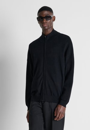 Uomo con maglione nero con zip, pantaloni neri e grandi occhiali da sole neri, in piedi davanti a uno sfondo chiaro e uniforme.