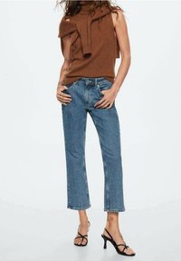 Top marrón sin mangas con un detalle de nudo, combinado con jeans de denim azul de tiro alto, cortados a la altura del tobillo, y sandalias negras de tacón alto con tiras.