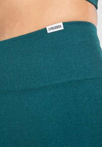 Teal geribbelde leggings met een brede, rekbare tailleband. De tailleband heeft een klein wit label met daarop "SMILODDXX" gedrukt.