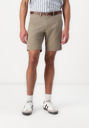 Pantaloni corti beige a taglio diritto, con passanti per cintura e risvolti, abbinati a sneakers bianche con strisce nere e dettagli grigi.