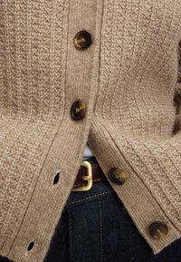 Cárdigan de punto beige con tejido texturizado, grandes botones marrones y una cinturilla con una hebilla dorada. Primer plano de los detalles y la costura.