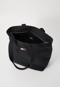 Svart lerret tote bag med to robuste håndtak, med glidelås og en innvendig etikett i rød, hvit og blå farger.