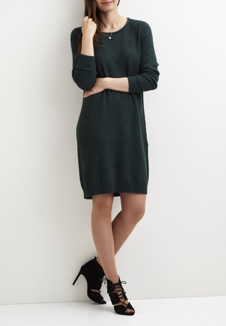 Robe en tricot vert foncé, dotée de manches longues, bords côtelés et ourlet, portée avec des chaussures à talons hauts en dentelle noires. Texture lisse, coupe décontractée.