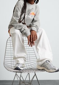 Felpa grigia con logo, pantaloni bianchi larghi e scarpe da ginnastica grigie. Seduto su una sedia di filo con le mani poggiate sulle ginocchia.