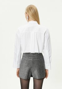 Weißes, knopfüberstehendes Hemd mit einem verkürzten Rücken, kombiniert mit schimmernden silbernen Shorts. Das Model trägt dunkle Strumpfhosen und große, abstrakte Ohrringe.