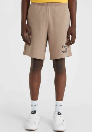 Beige Shorts mit elastischem Bund, Seitentaschen und dem aufgedruckten Text "The Future Surf Society" am linken Bein. Getragen mit weißen Sneakers und Socken.