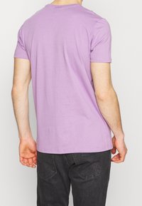 Krátký rukáv levandulového trička vyrobeného z bavlny, s kulatým výstřihem a hladkou texturou. Na sobě má tmavě šedé džíny, viděno zezadu.