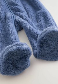 Un vêtement en polaire bleu présente une texture douce, de larges poignets et une fermeture éclair, conçu pour le confort et la chaleur. Les tissus semblent moelleux et accueillants.
