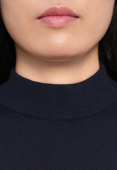 Tumši zila izspēlētā turtlenecks džemperis ar rievotu apkakli, izgatavots no mīkstas auduma. Tuvplāns, kas parāda gludu ādu un lūpas.