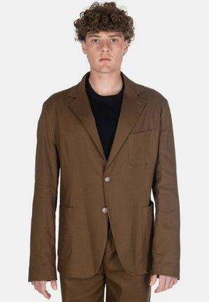 Giovane uomo con capelli ricci che indossa un blazer marrone a due bottoni e pantaloni abbinati sopra una camicia nera, in piedi davanti a uno sfondo semplice.