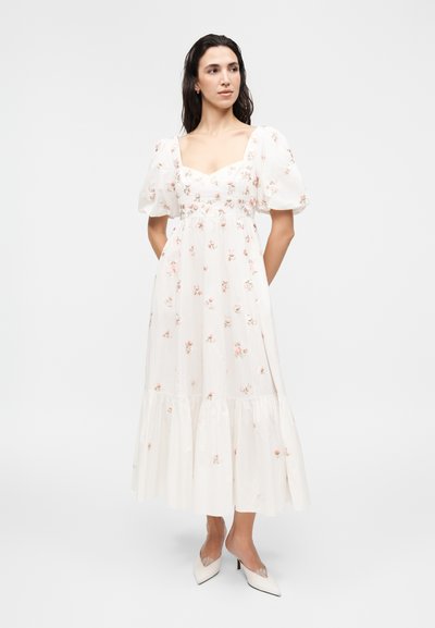 Γυναίκα που φοράει λευκό midi φόρεμα με φουσκωτά μανίκια και ροζ λουλουδένια σχέδια, συνδυασμένο με λευκές μυτερές γόβες, στέκεται με τα χέρια πίσω στην πλάτη.