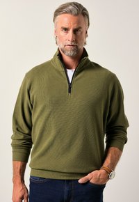 Sudadera de color verde oliva con cremallera, fabricada con un tejido texturizado, puños acanalados y un cuello alto. Combinada con jeans azul oscuro y un reloj marrón.