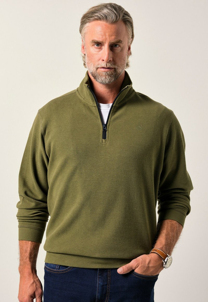Sudadera de color verde oliva con cremallera, fabricada con un tejido texturizado, puños acanalados y un cuello alto. Combinada con jeans azul oscuro y un reloj marrón.