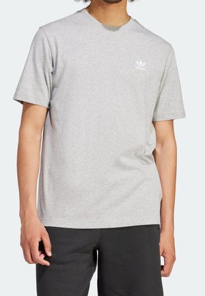 Mężczyzna w jasnoszarej koszulce Adidas z logo na piersi i czarnych spodniach, stojący na jednolitym tle.