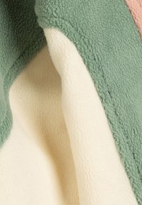 Coperta in pile con un'esterno verde testurizzato e un sottopancia morbido color crema, caratterizzata da un sottile design a blocchi di colore.