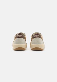 Zapatillas con parte superior de ante beige, acentos de purpurina dorada y parches de logo marrón. Suelas de goma planas con detalles texturizados. Vista trasera.