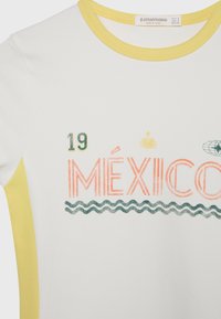 Chemise blanche avec bordure jaune, texte « MEXICO » en orange, lignes ondulées vertes en dessous, numéro 19 et un graphique de globe près de l'encolure.