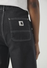 Μαύρο τζιν παντελόνι με πίσω τσέπη που έχει ετικέτα λογότυπου Carhartt σε γκρι και λεπτομέρειες με λευκή ραφή. Λείο ύφασμα, κανονική εφαρμογή.