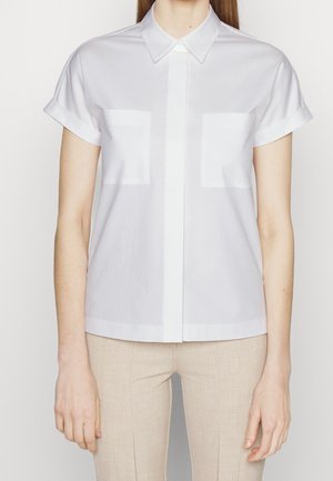 Kobieta ubrana w białą, krótkorękawową koszulę na guziki z dwoma kieszeniami na piersi oraz beżowe spodnie na jednolitym tle.