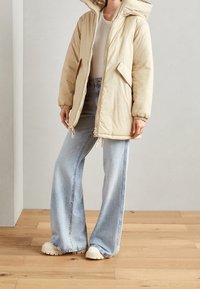 Chaqueta puffy beige con una gran capucha, cierre delantero y puños elásticos. Combinada con jeans de pierna ancha azul y zapatillas de plataforma blancas. Fondo de suelo de madera.